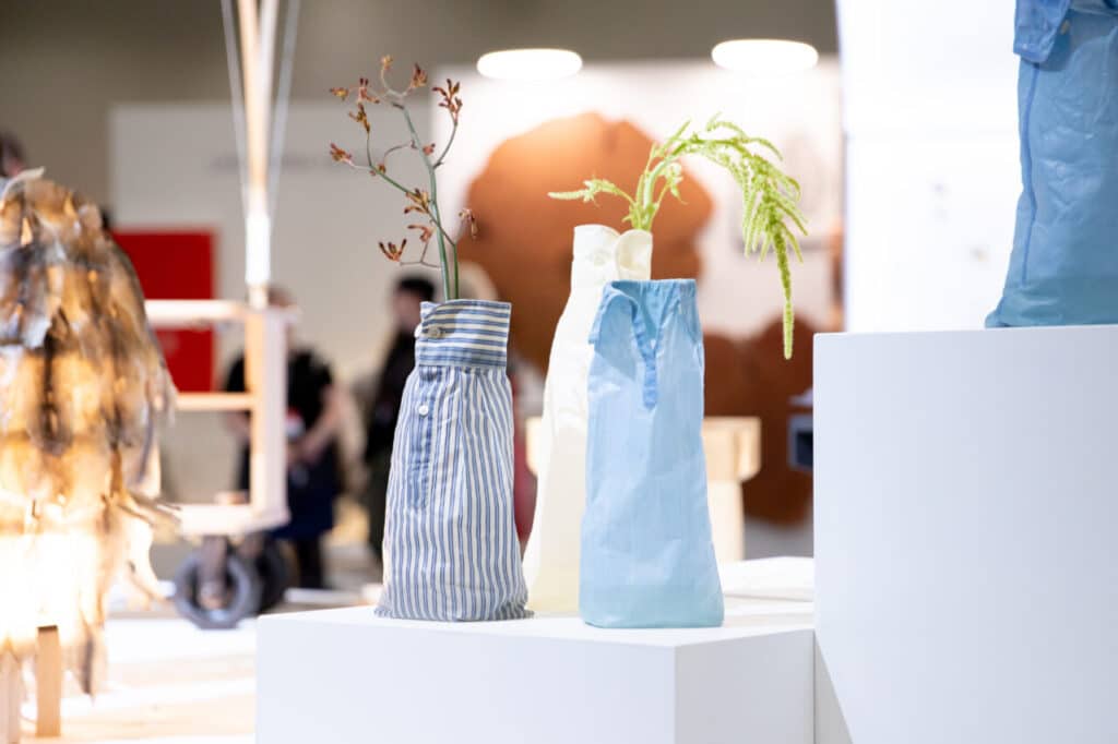 Three fabric-covered vases in striped, blue, and white materials, each holding a different type of flower, displayed on a pedestal.