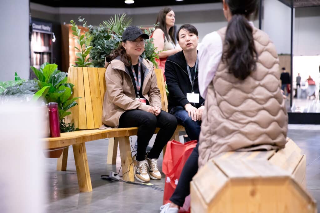 Two people sitting on a wooden bench planter, talking with a third person standing in front of them.