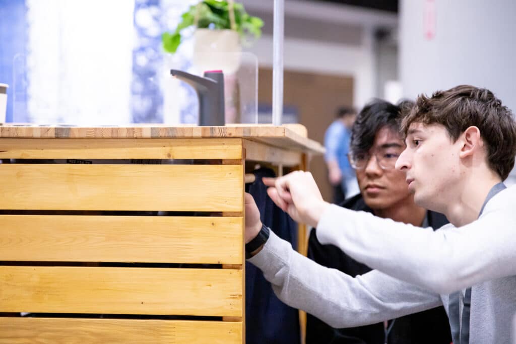 Close-up of two people crouched beside a wooden structure, examining and discussing its details.