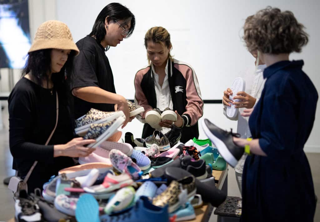 Participants examine and compare shoes laid out on a table, picking up pairs and discussing them in a group setting.