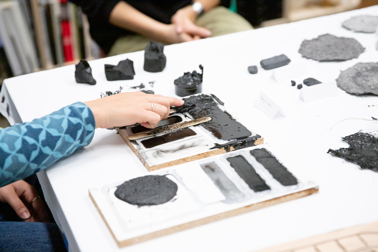 A white table with samples of materials made from charred wood, all black or grey. A hand points to one of the samples.