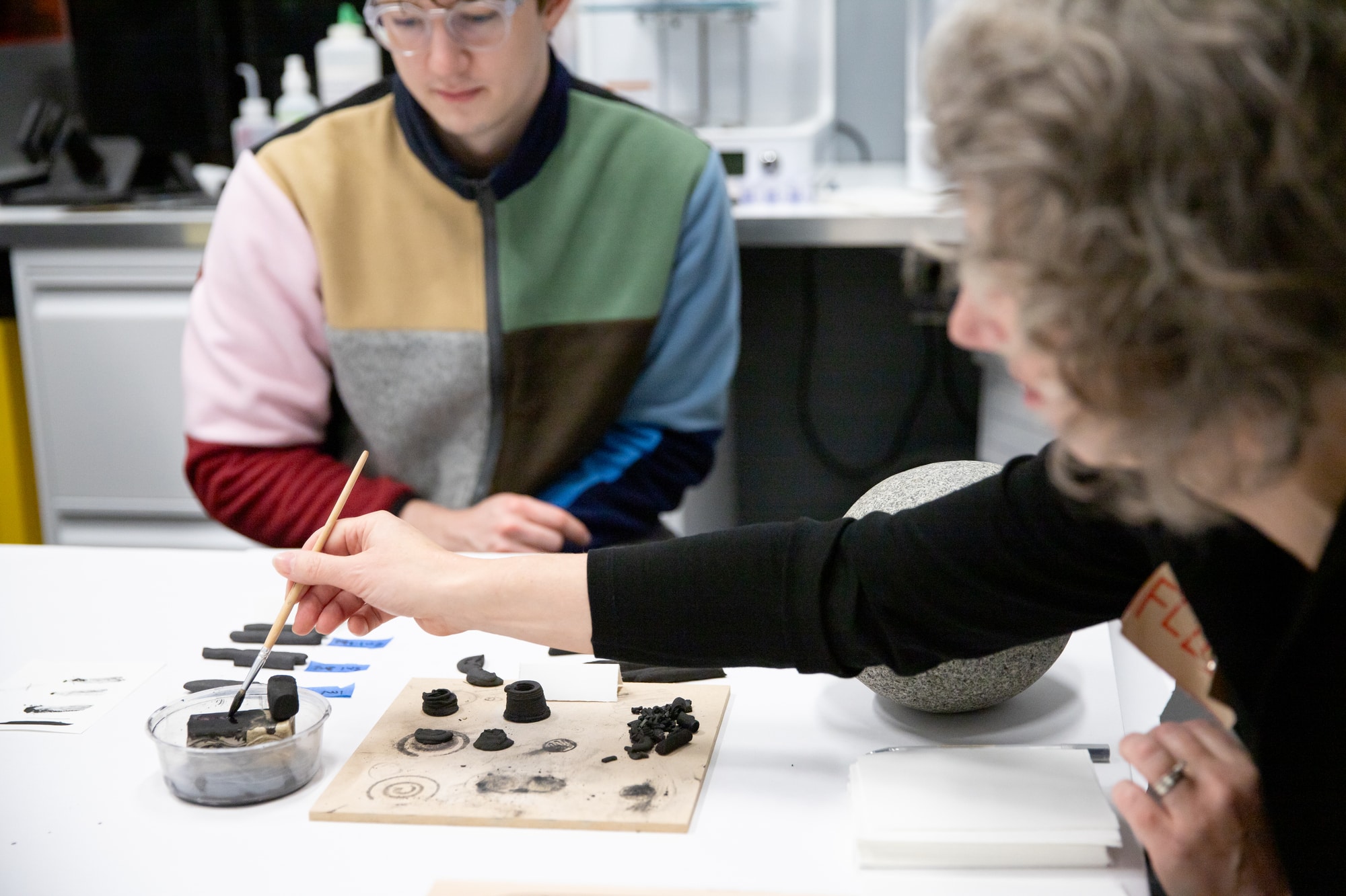 A person paints small charcoal test pieces on a wooden board with a brush while another student watches.