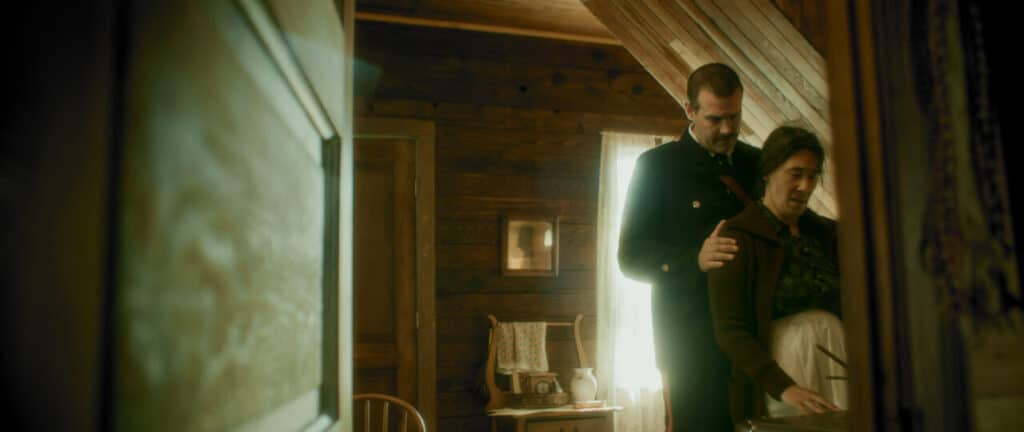 Inside a wooden house, an RCMP officer stands behind an older adult, resting their hands on the adult’s shoulders. The adult looks downward, holding a blanket, while soft light filters through lace curtains in the background.