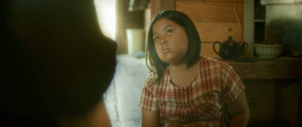 A child wearing a red plaid dress sits in a rustic wooden room, looking upward with a serious expression. Light streams through a doorway behind them, and a teapot and bowl rest on a shelf.