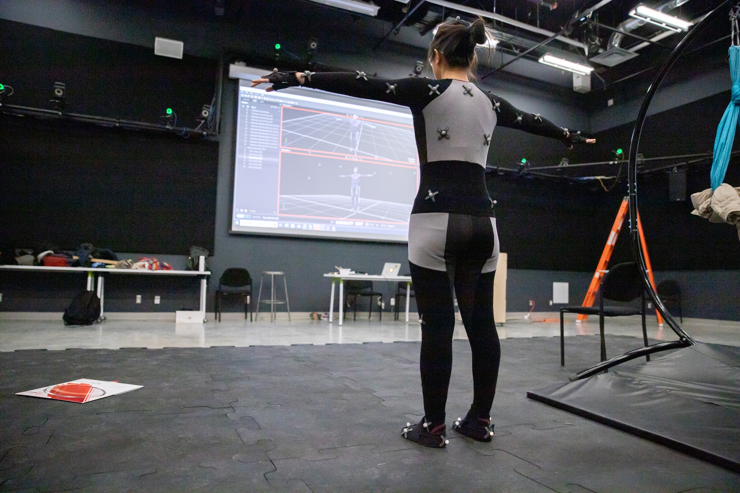 A student stands with arms extended, wearing a black motion capture suit in a large black-walled room with a screen.
