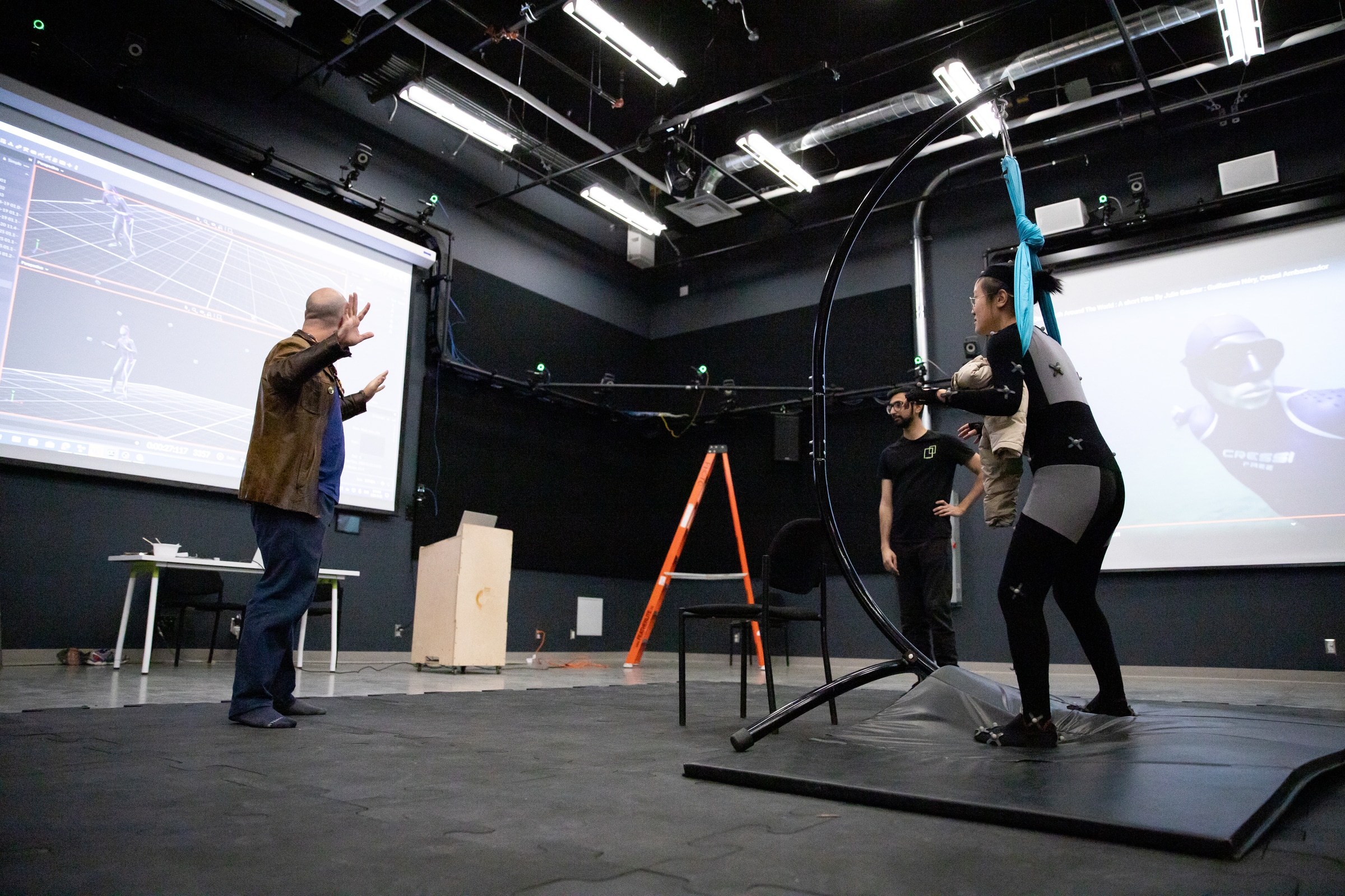 Motion capture studio with a person in a sensor suit suspended in a rig, guided by an instructor while another observer stands nearby, with 3D skeletal animation projected on a large screen.