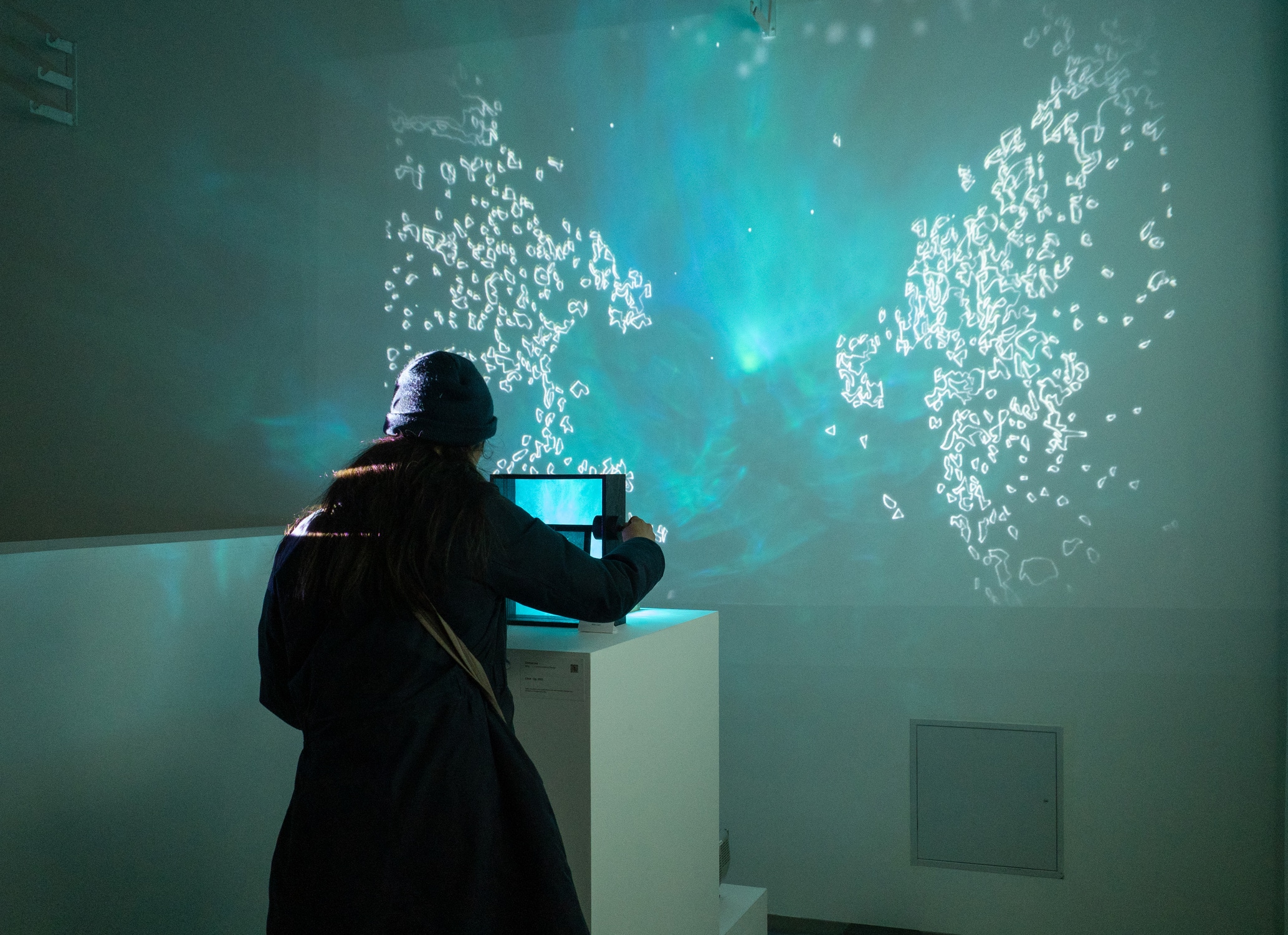 A person in a dark coat and knit hat interacts with an illuminated installation in a dim gallery. They stand before a glowing blue-green projection on the wall that resembles rippling water with clusters of light outlines, creating an immersive effect.