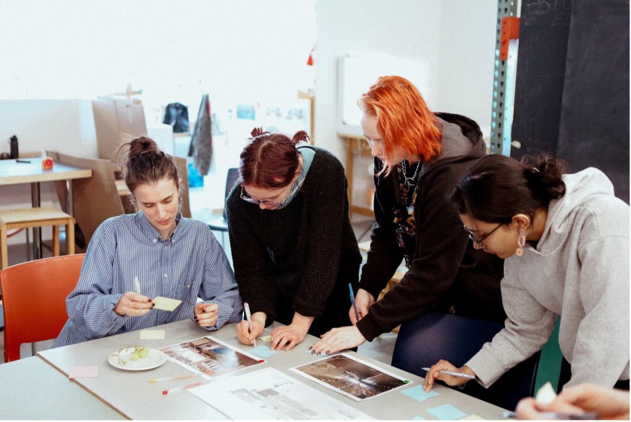 Students collaborate at a table, writing notes on paper and sticky notes as they develop ideas in a classroom workshop.