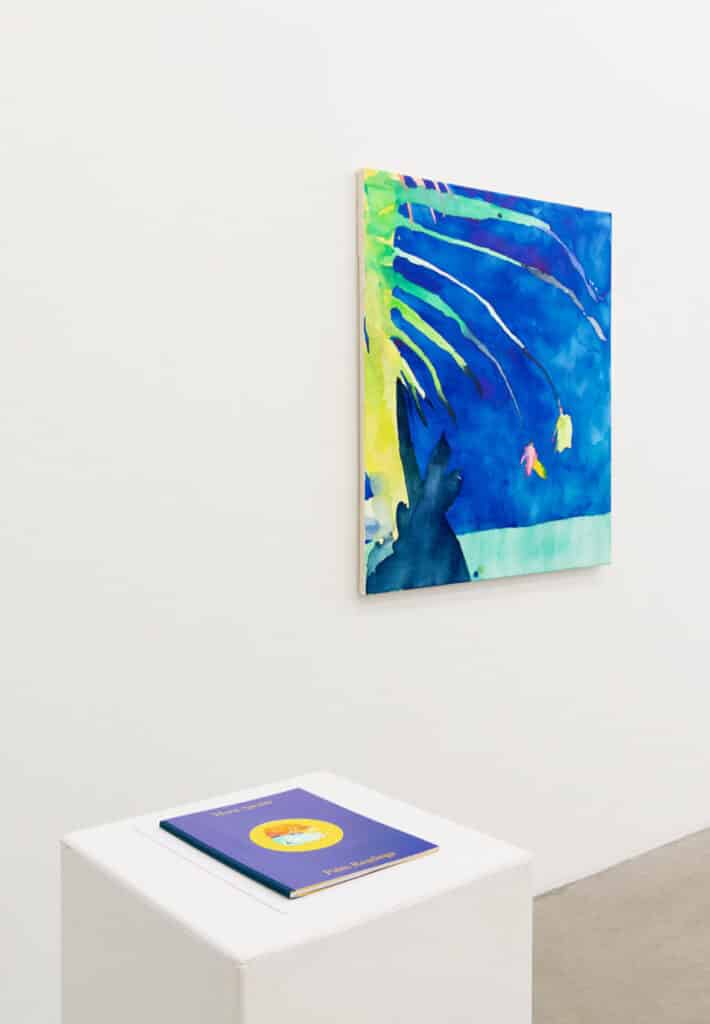 A bright abstract painting in blue, green and yellow on a gallery wall, with a pedestal below displaying a blue-covered book titled Palm Readings.