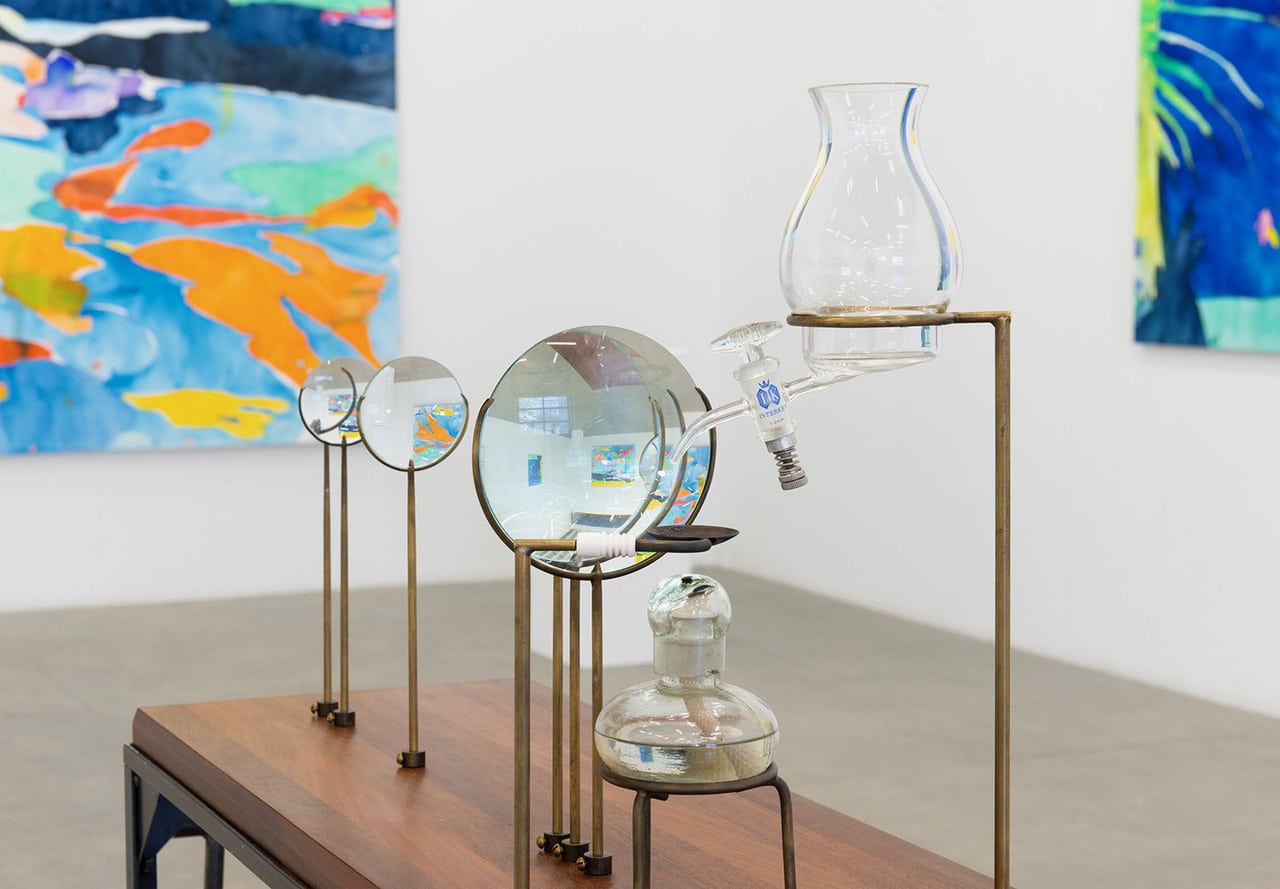 Glass vessels and magnifying lenses arranged on a wooden table with colourful abstract paintings visible in the background.