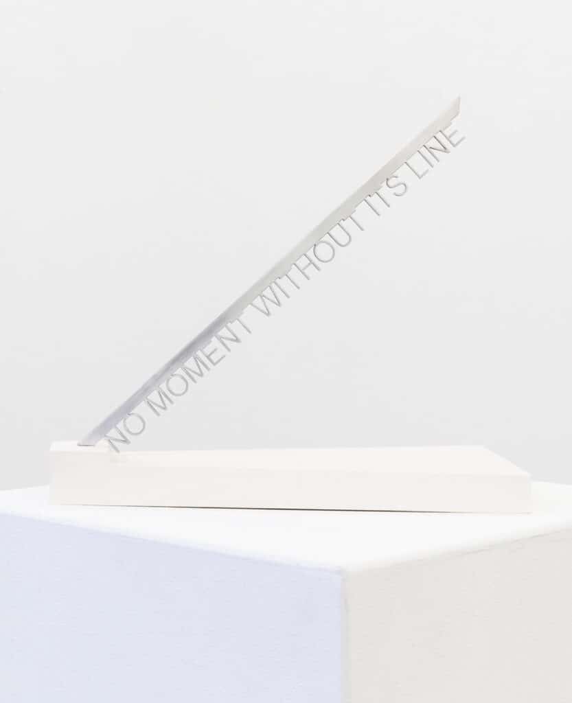 A white pedestal holding a diagonal white bar inscribed with the phrase “No Moment Without Its Line.”