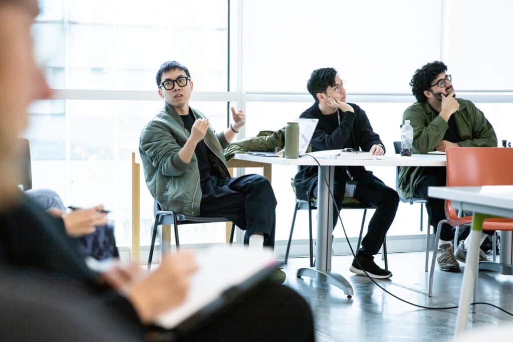 A student gestures while speaking during a classroom discussion. Two classmates beside him listen attentively, one resting his chin on his hand.