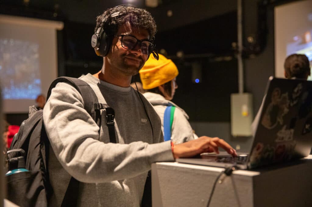 Person wearing headphones and a backpack smiling while using a laptop in a darkened room with projections on the wall.