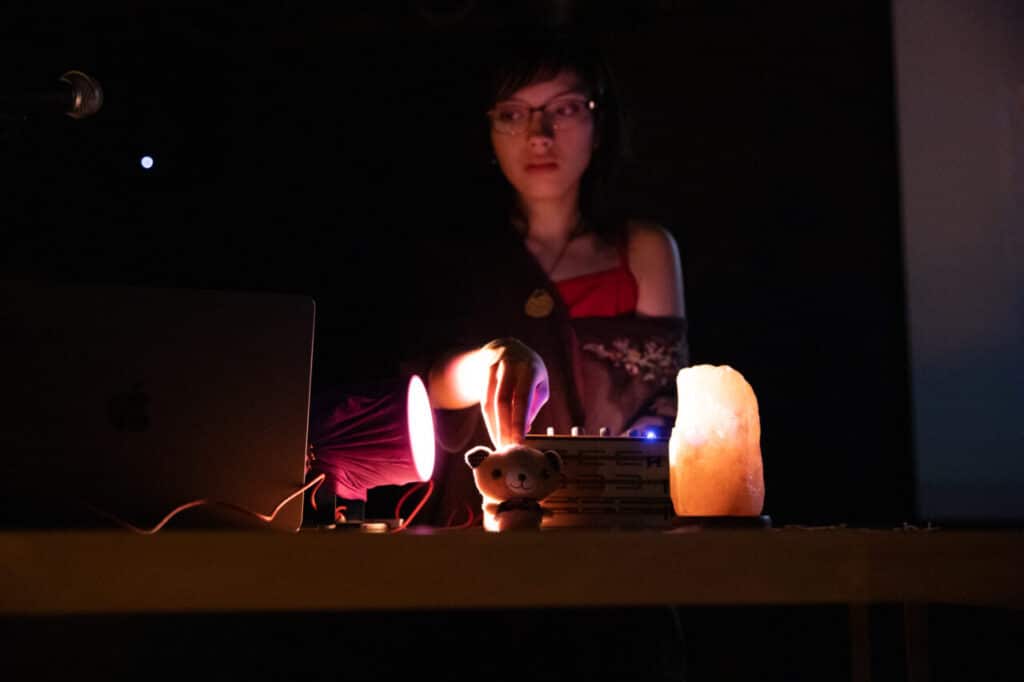 Person standing behind a table in a dimly lit space, adjusting sound equipment surrounded by a glowing lamp and small toy figure.