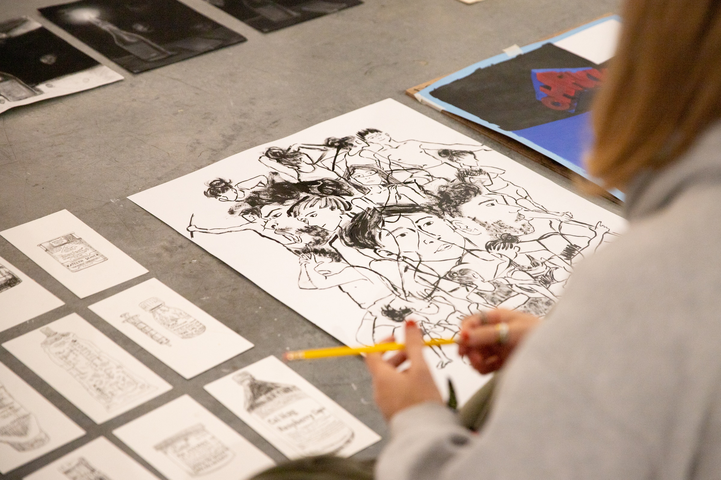 Student sketching with a pencil on paper featuring expressive black ink drawings of faces, surrounded by smaller ink illustrations of objects.