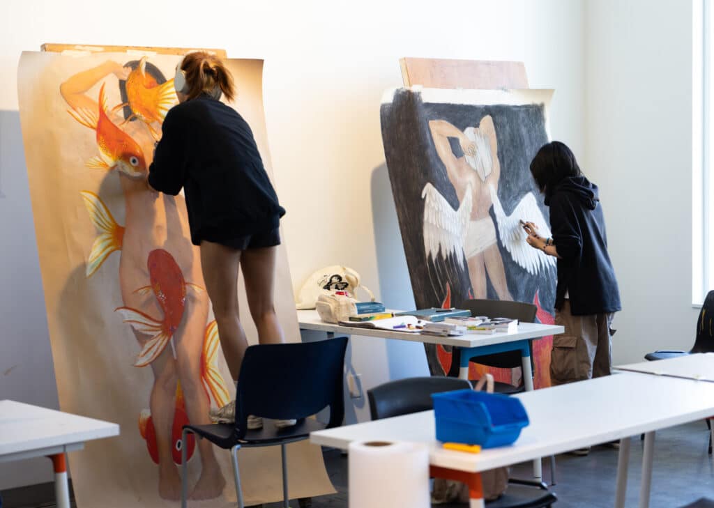 Two students painting large figure drawings — One student stands on a chair painting a large drawing of a nude figure surrounded by goldfish, while another student works on a drawing of a winged figure against a dark background.
