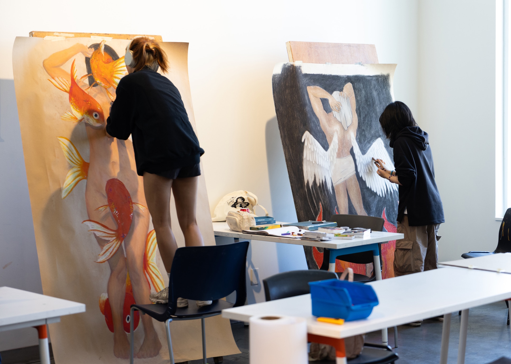 Two students painting large figure drawings — One student stands on a chair painting a large drawing of a nude figure surrounded by goldfish, while another student works on a drawing of a winged figure against a dark background.
