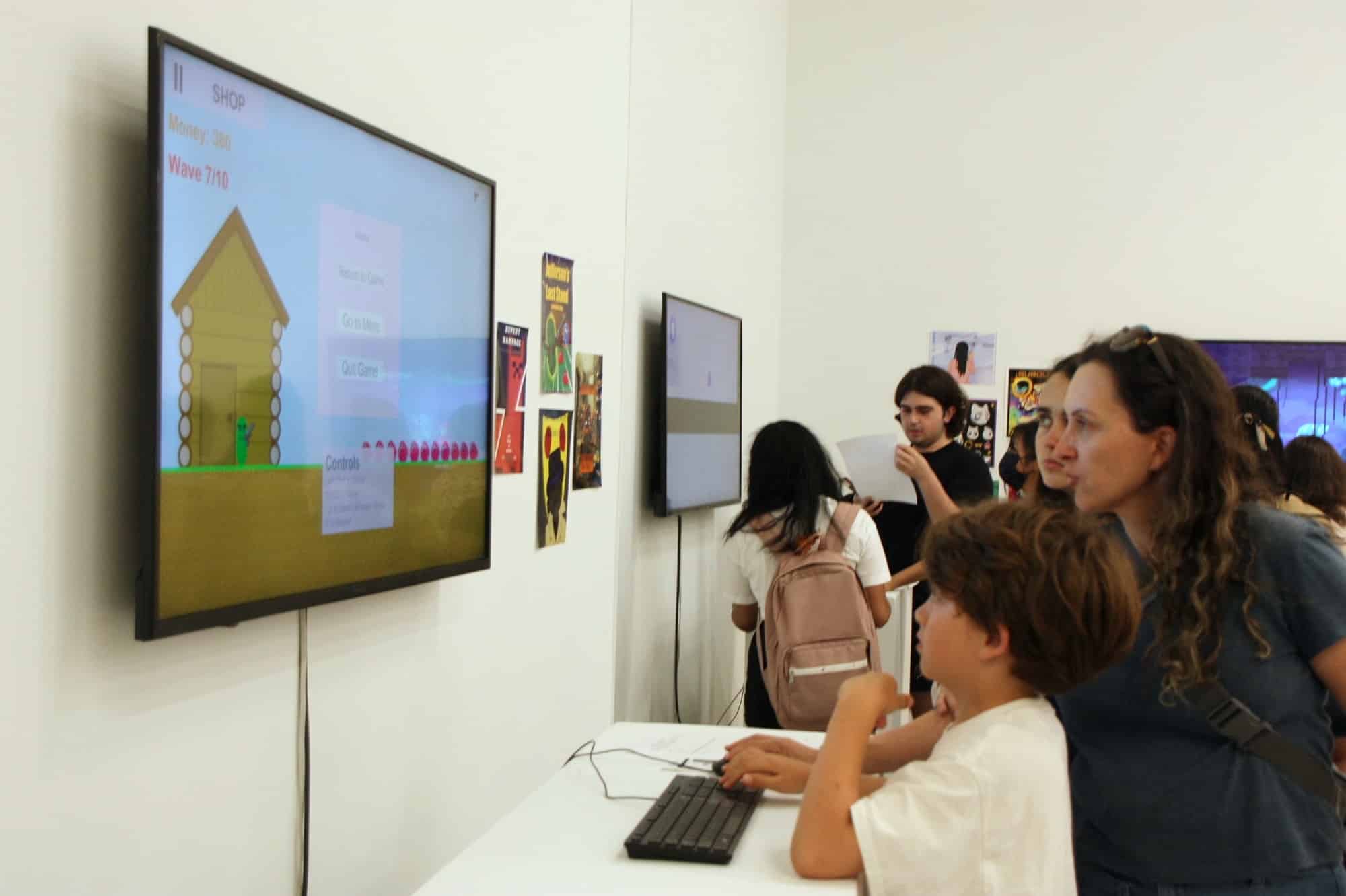 Young boy playing a student-designed computer game on a wall-mounted screen while an adult and other visitors look on.