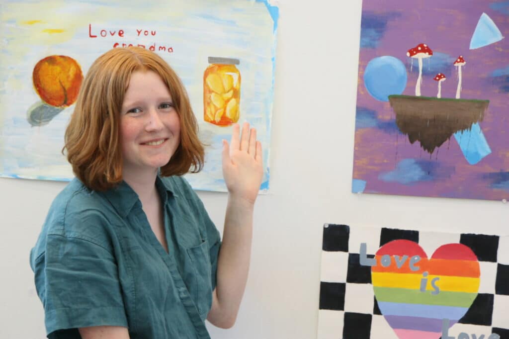 Smiling teen standing beside their artwork, including paintings of fruit, jars, mushrooms, and a rainbow heart with text reading “Love is Love.”