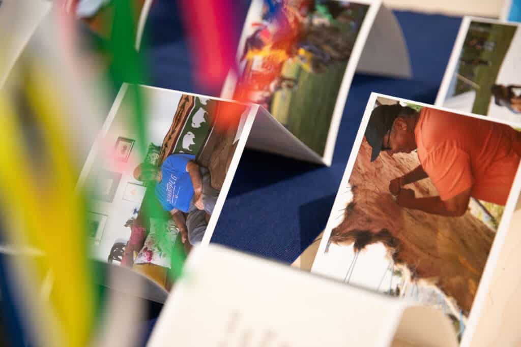 Close-up view of printed photographs displayed on a table, featuring candid images of people engaged in daily activities, with colorful paper strips slightly obscuring the view.