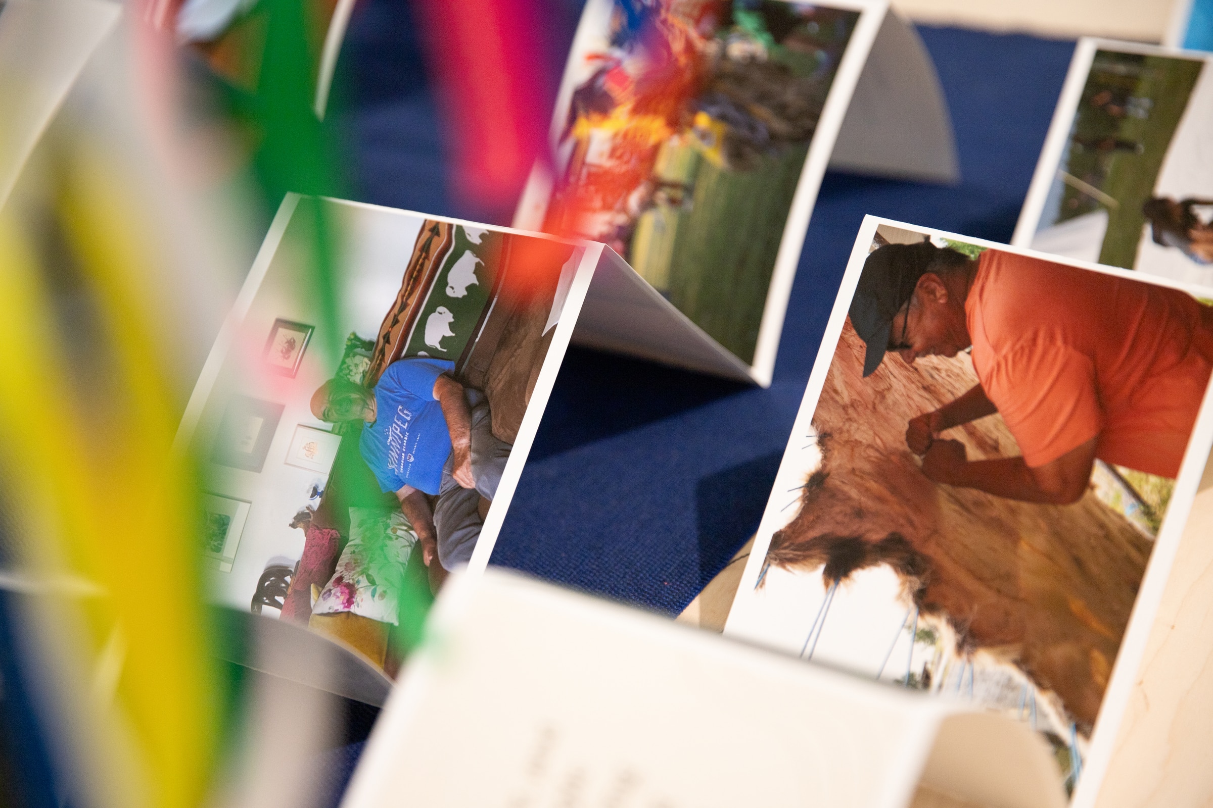 Close-up view of printed photographs displayed on a table, featuring candid images of people engaged in daily activities, with colorful paper strips slightly obscuring the view.