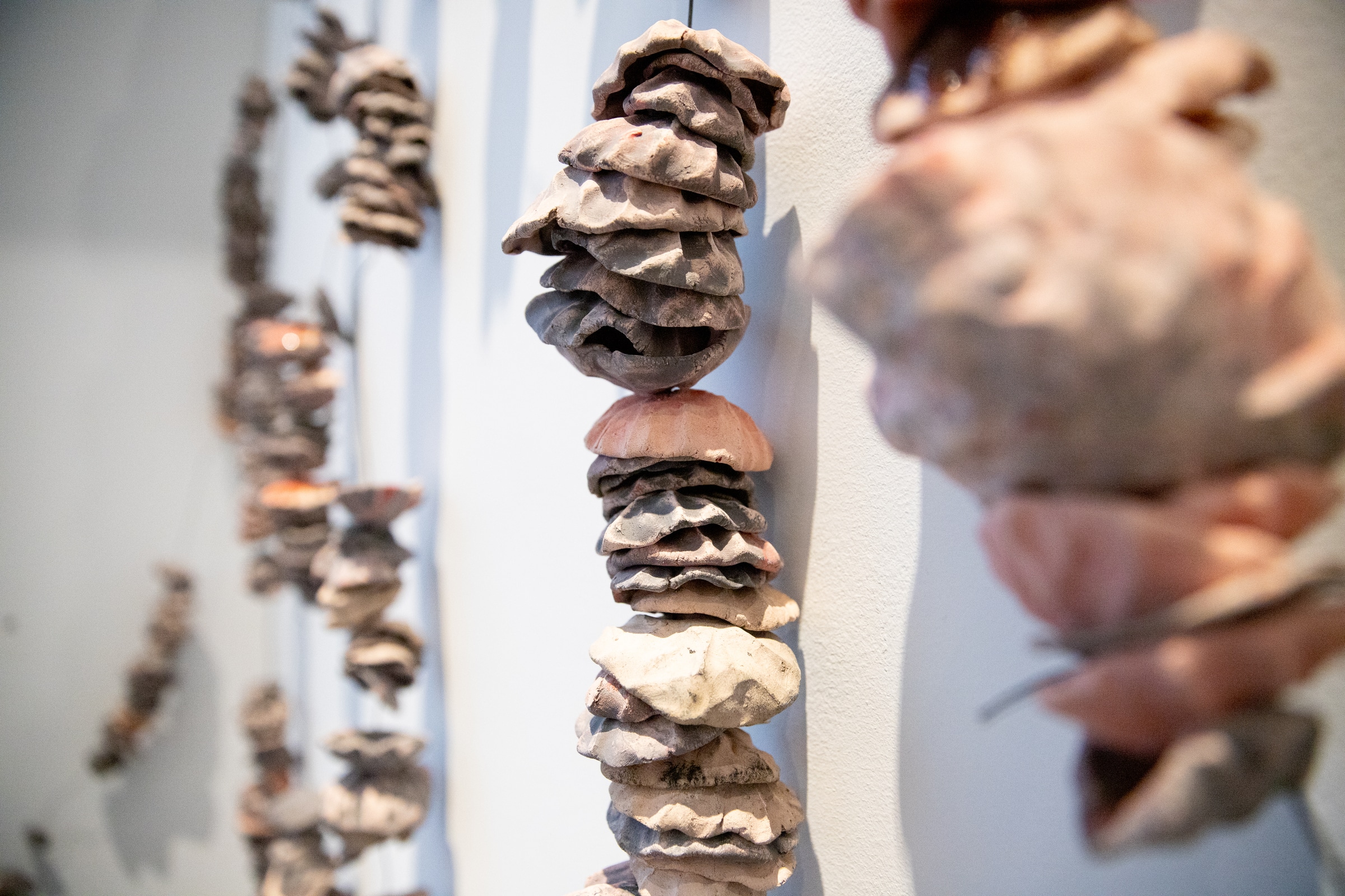 Close-up view of a vertical installation of stacked, organic-shaped ceramic forms strung together and hanging against a white wall, with similar strands receding into the background.