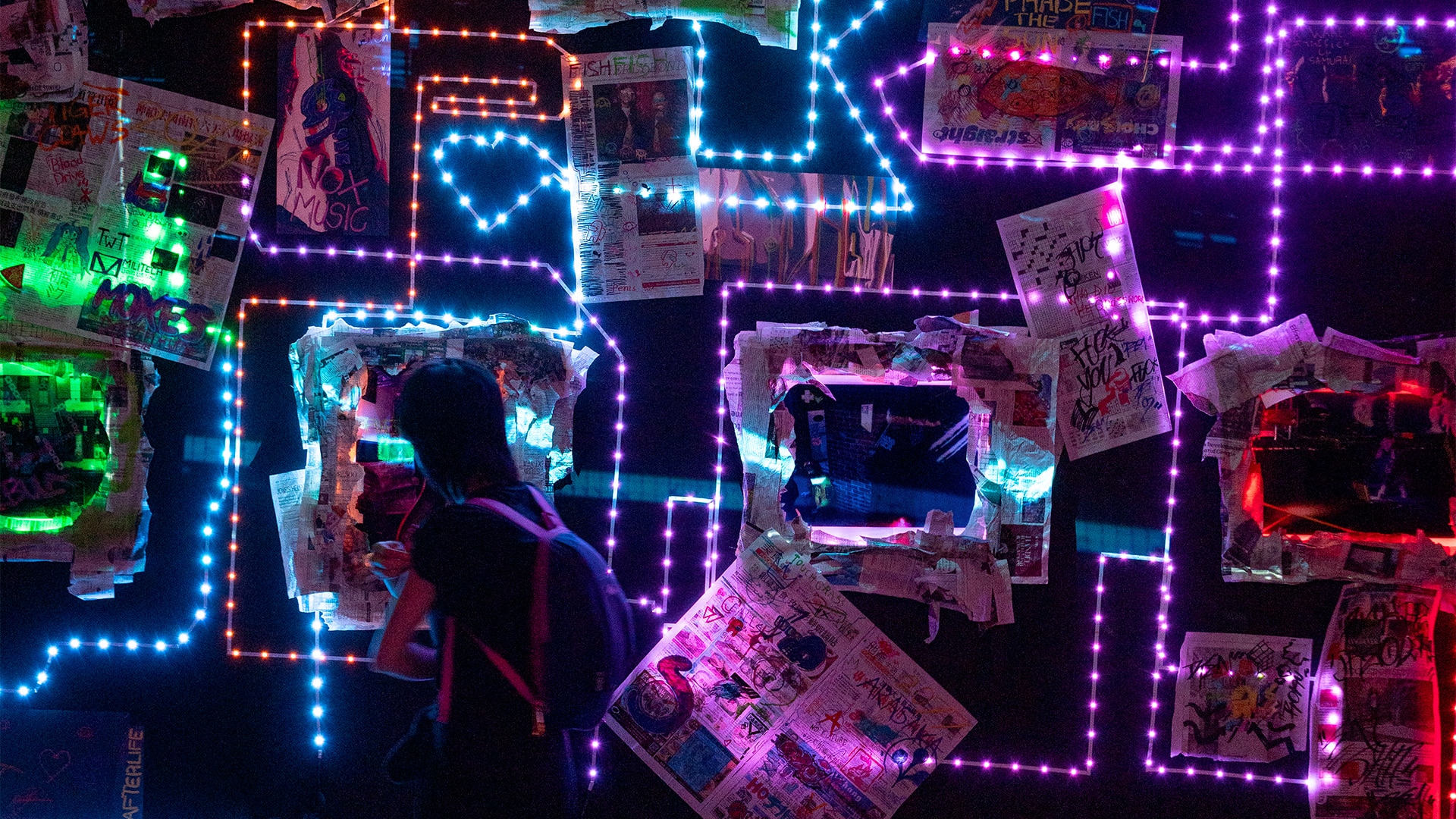 A viewer observes a neon-lit wall installation of photos, newspaper clippings, and LED outlines at the 2024 Ghost Images – Photography and Trace exhibition.