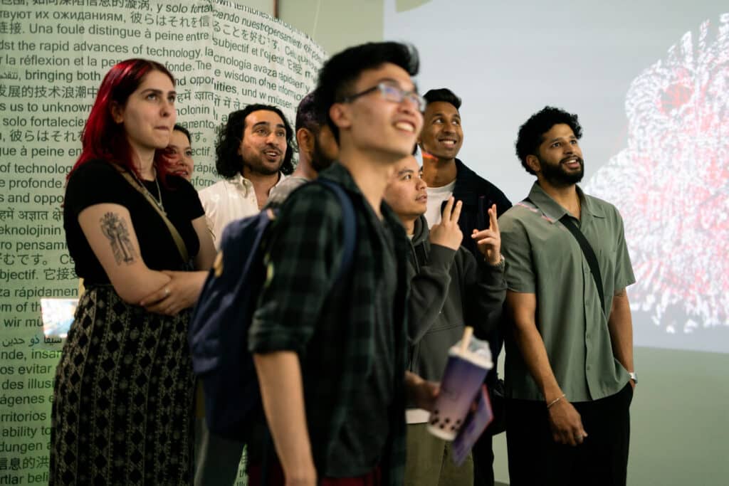 A group of students stand together, smiling and looking at a large projected screen with text in multiple languages.