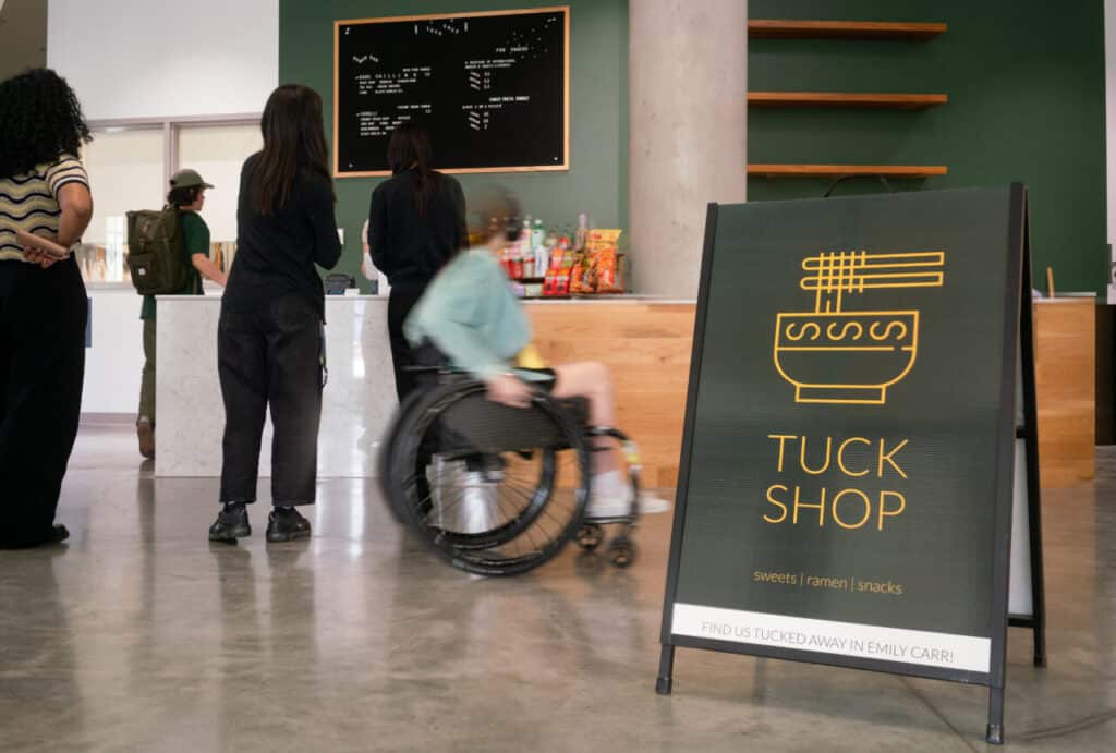 Students line up at the Tuck Shop counter at Emily Carr University, with a sign in the foreground reading “Tuck Shop – sweets, ramen, snacks.”