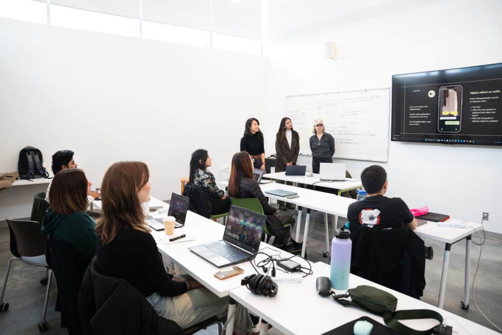 Group of students seated at desks with laptops watching a presentation projected on a screen at the front of the classroom.
