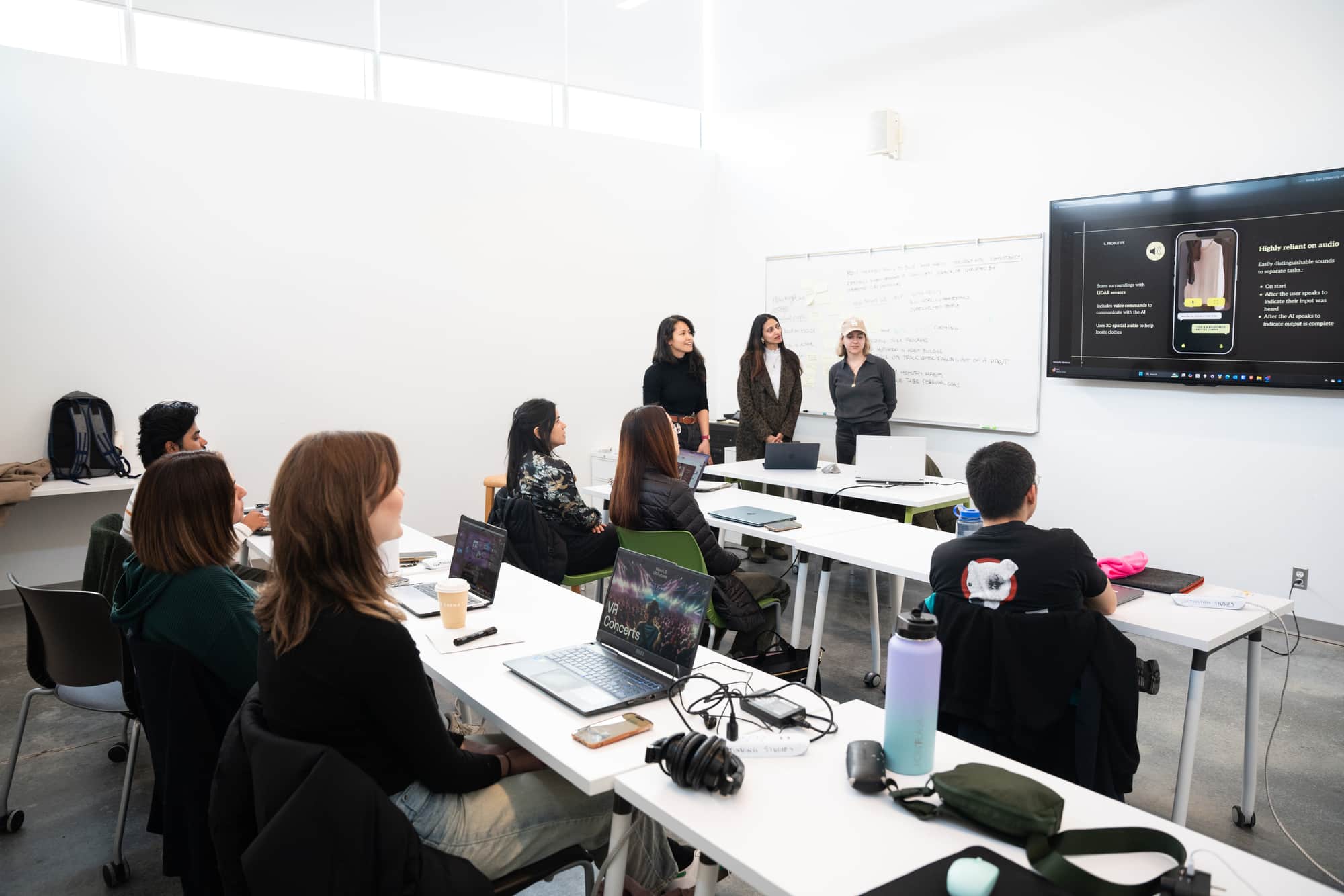 Group of students seated at desks with laptops watching a presentation projected on a screen at the front of the classroom.