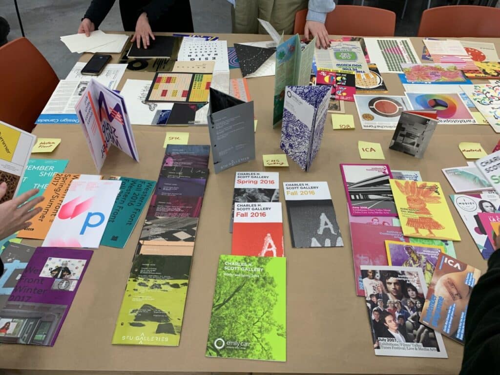 Table covered with a wide variety of colourful printed booklets, brochures and folded publications, with people’s hands reaching in to examine them.