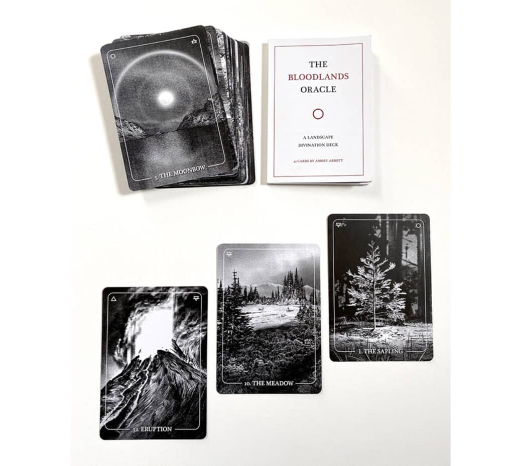 Oracle card deck titled “The Bloodlands Oracle” shown with cards spread out, including images of a volcano eruption, meadow, sapling and moonbow