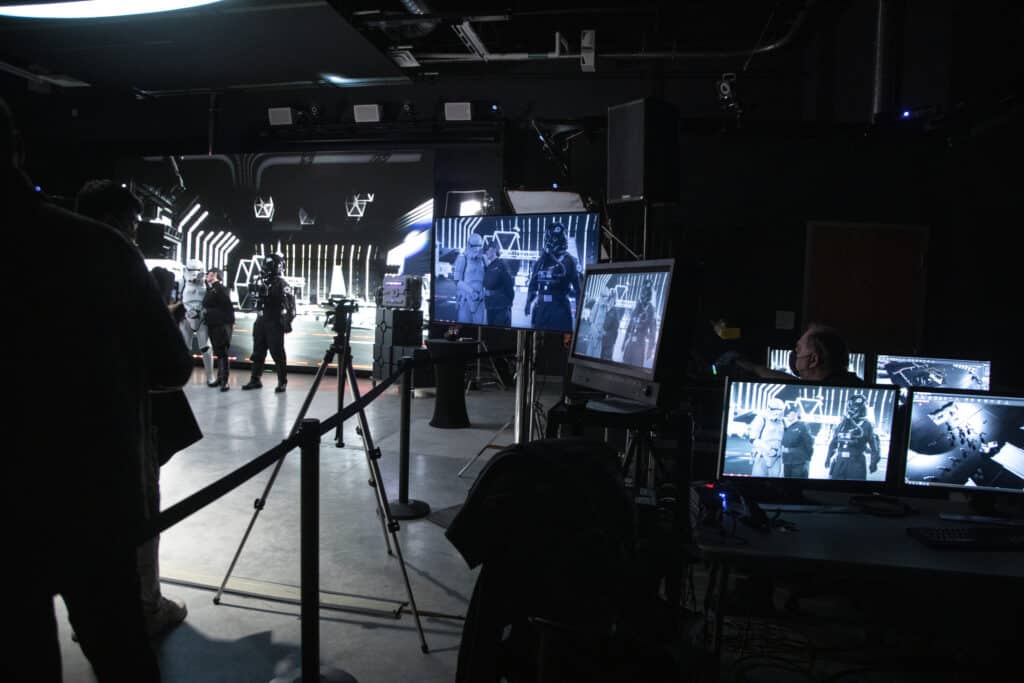 A behind-the-scenes view of a virtual production studio, with large LED screens displaying a futuristic sci-fi scene. Several people in costumes, including stormtroopers, stand in front of the screen while multiple monitors show live camera feeds of the set.