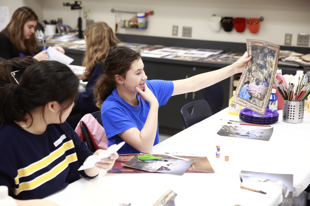 A group of young students sit around a long white table in an art classroom. One student in a bright blue shirt smiles and holds up a picture with a decorative border, showing a scene of people in a garden. Another student beside her looks at a paper cut-out while the table is covered with scissors, glue sticks, brushes, and art supplies.