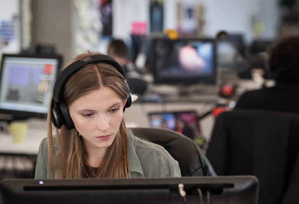 Student wearing headphones and working at a computer in a busy design studio environment.