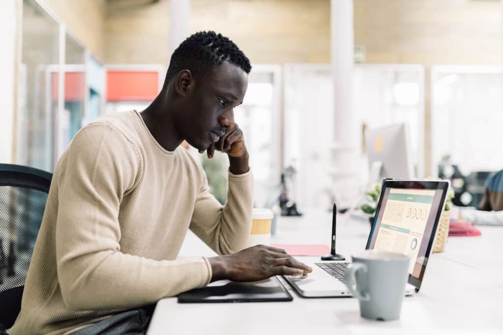 Person in a beige sweater focused on a laptop at a shared workspace, with charts visible on the screen.