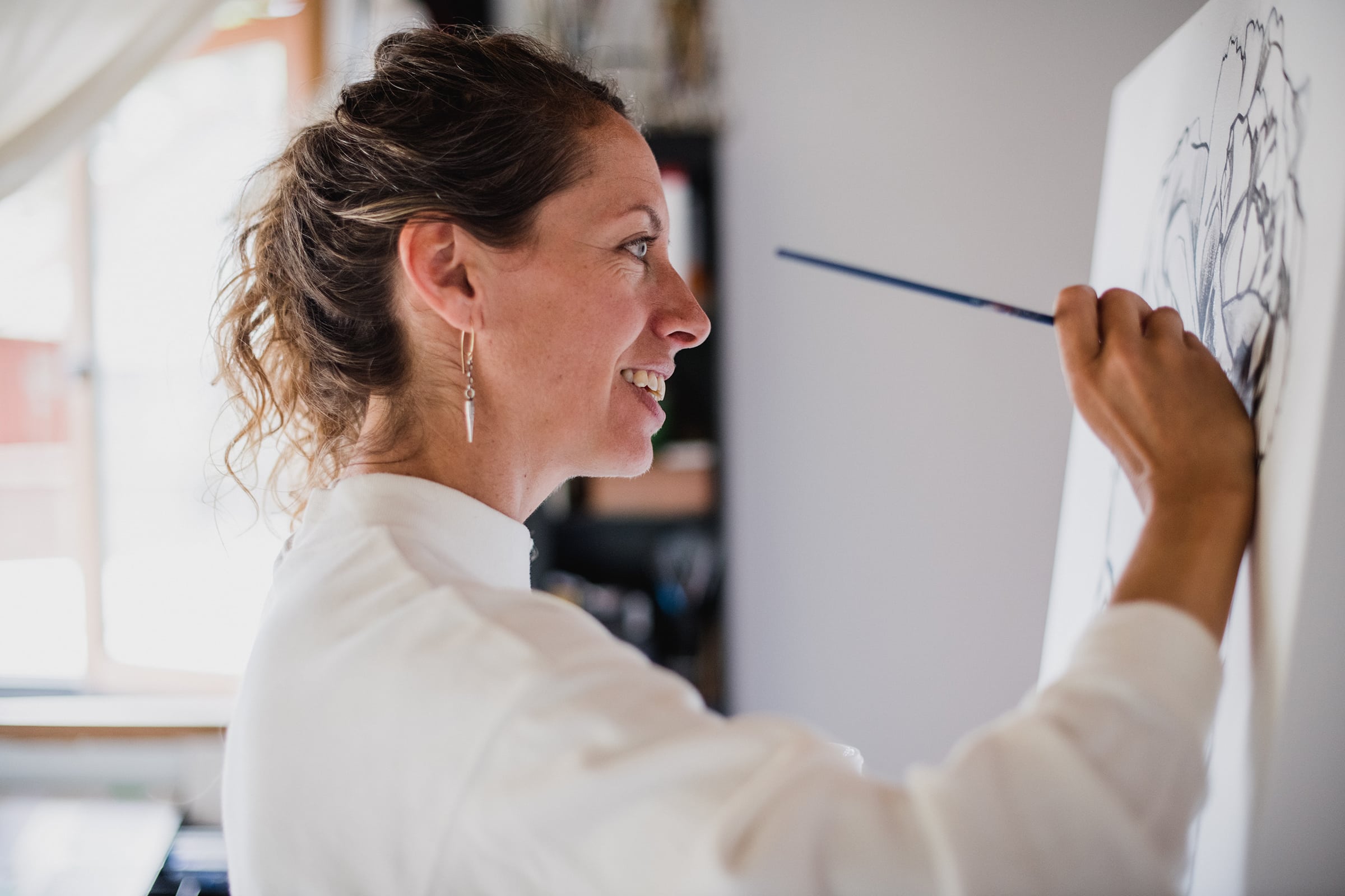 Smiling artist painting a detailed black-and-white flower illustration on a canvas indoors.