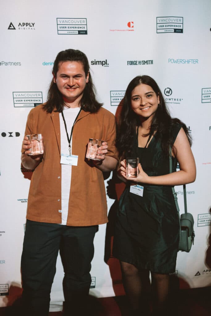Two people standing in front of a sponsor backdrop at the Vancouver User Experience Awards, smiling and holding glass awards.