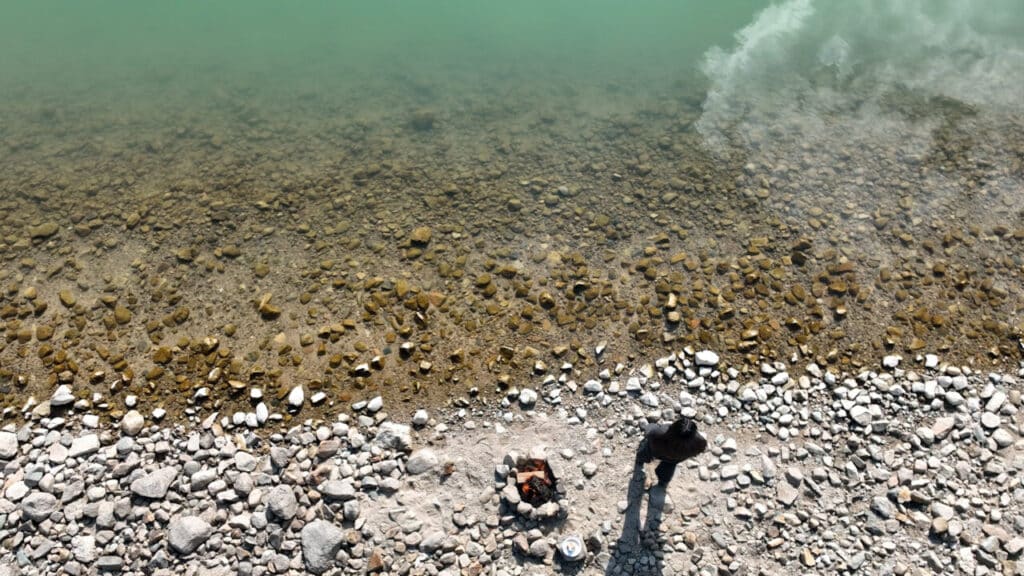 Overhead view of a rocky shoreline where a person in dark clothing stands by a small fire pit near green water.