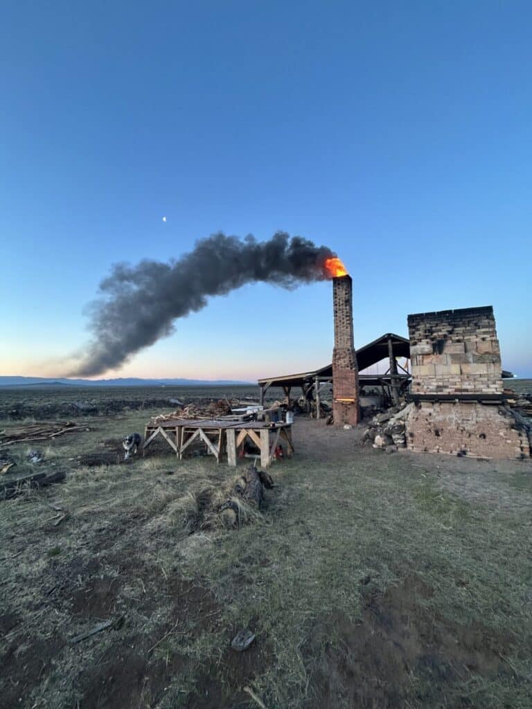 Brick kiln with fire and black smoke rising from the chimney in a rural landscape at dusk.