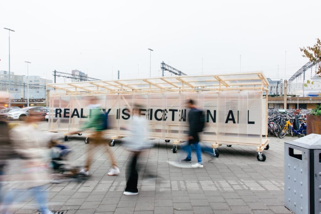 Street view of a translucent pavilion on wheels with the words “Reality is Fictional” visible, while blurred pedestrians walk past.