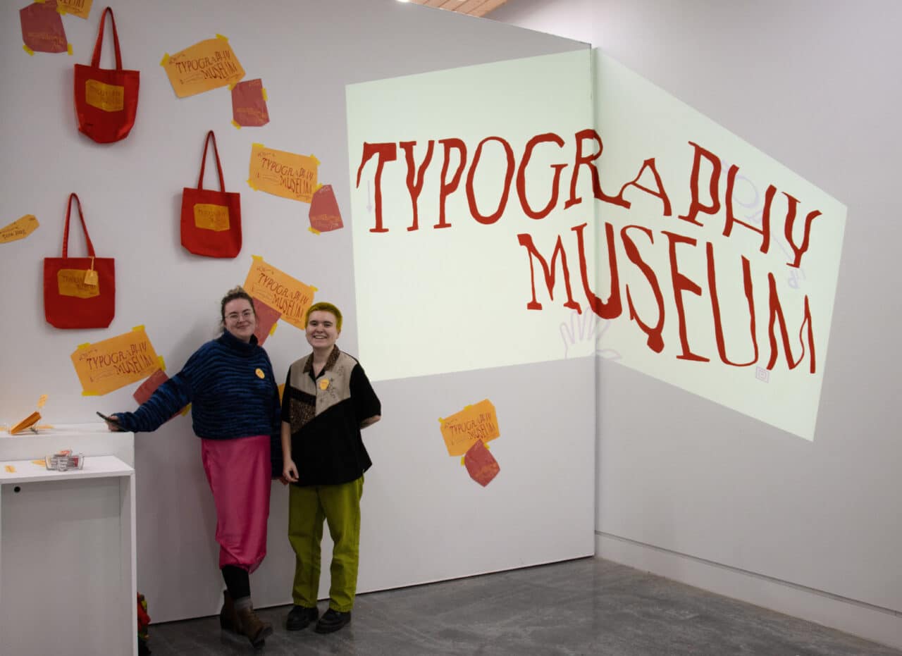 Two students stand smiling in front of a wall projection reading “Typography Museum,” surrounded by bright red tote bags and posters.