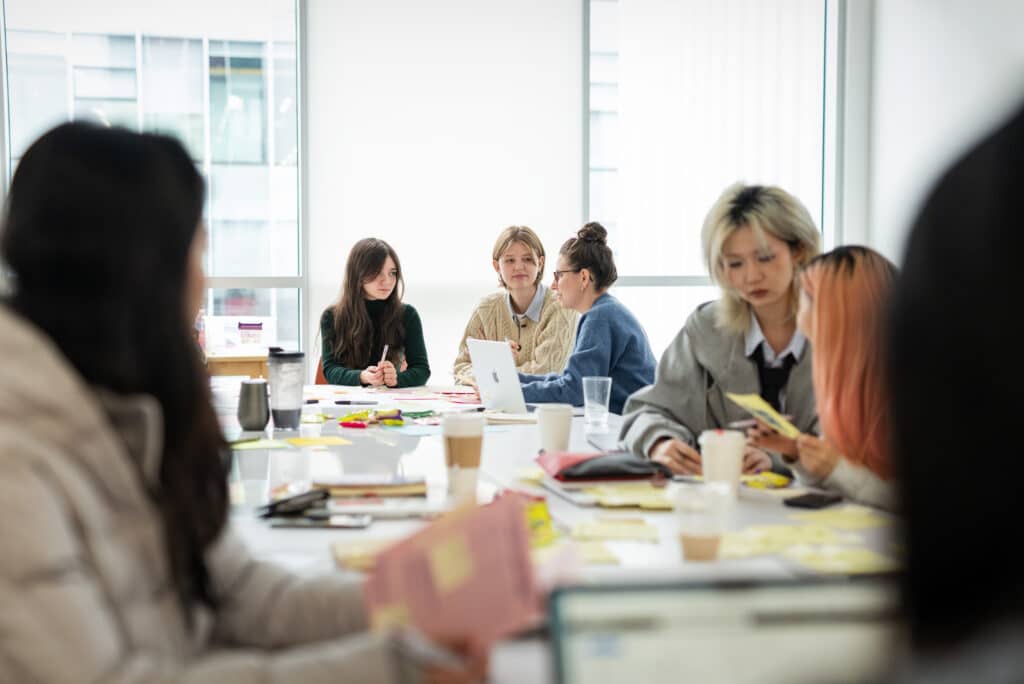 Students gather around tables covered with sticky notes, laptops, and papers, collaborating in a bright classroom.