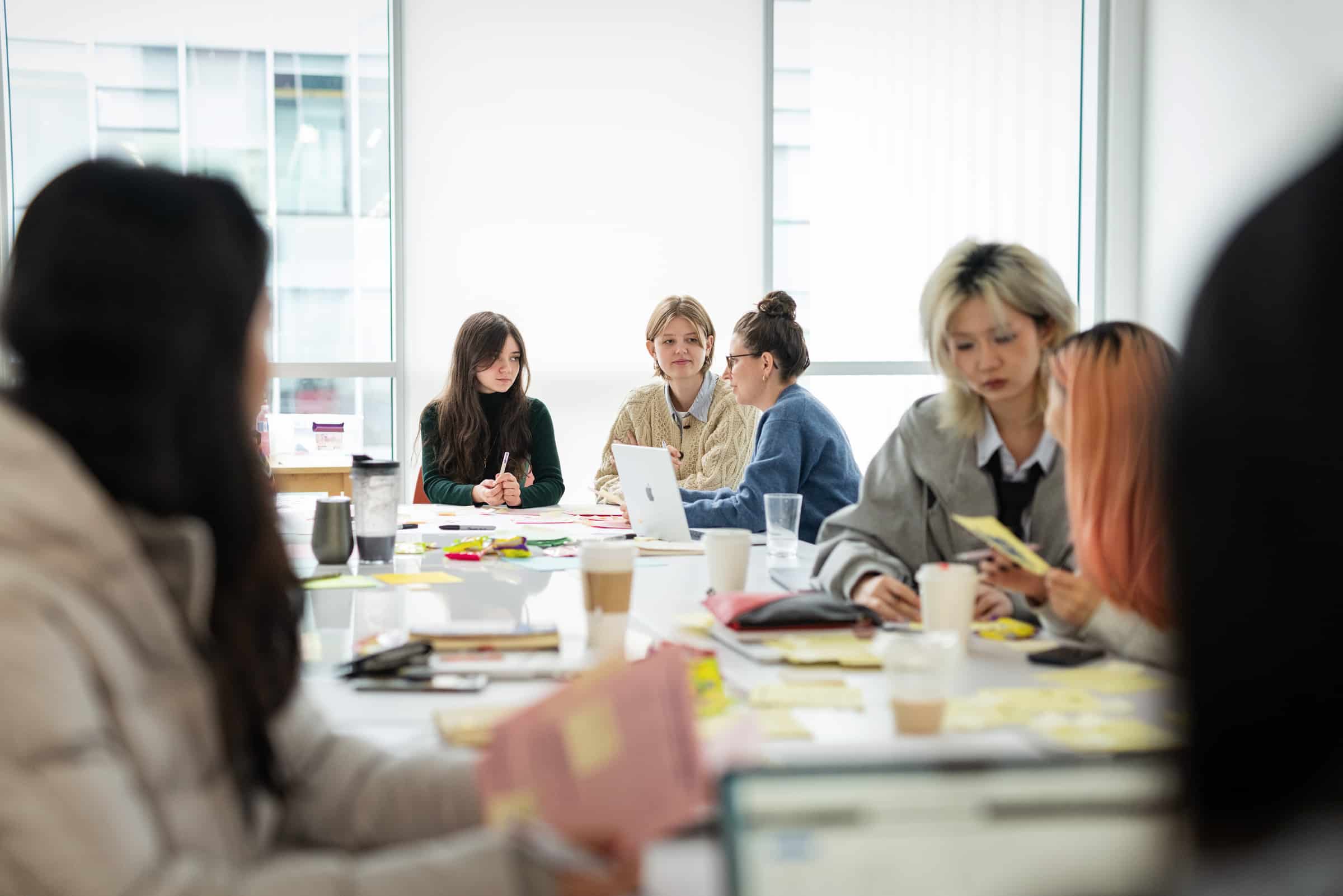 Students gather around tables covered with sticky notes, laptops, and papers, collaborating in a bright classroom.