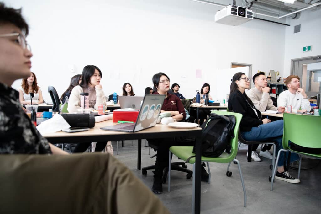 A group of students sit in a classroom, focused on a presentation. Laptops, notebooks, and drinks are on the tables.
