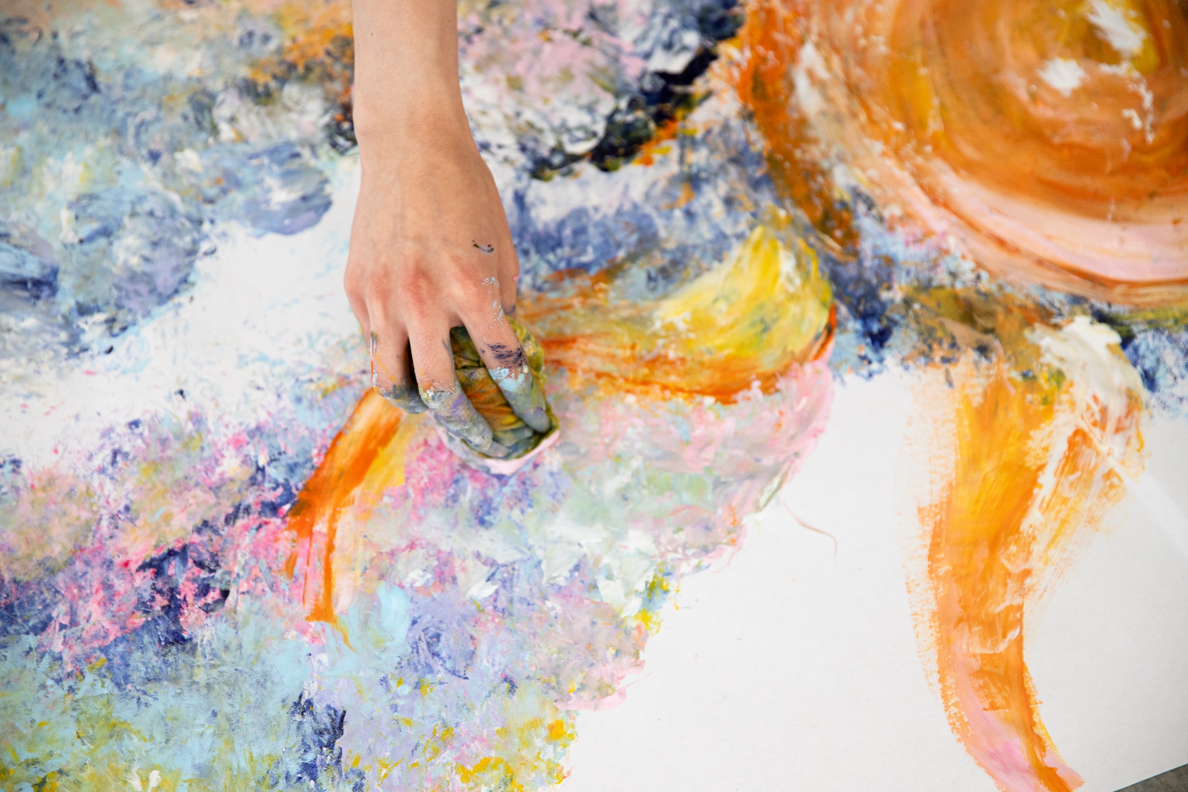 Close-up of a student kneeling on the floor, painting with bright yellow and orange acrylics on a large sheet of paper. Swirls of vibrant colors—pink, blue, yellow, and orange—create an abstract composition. Open paint containers surround the work.