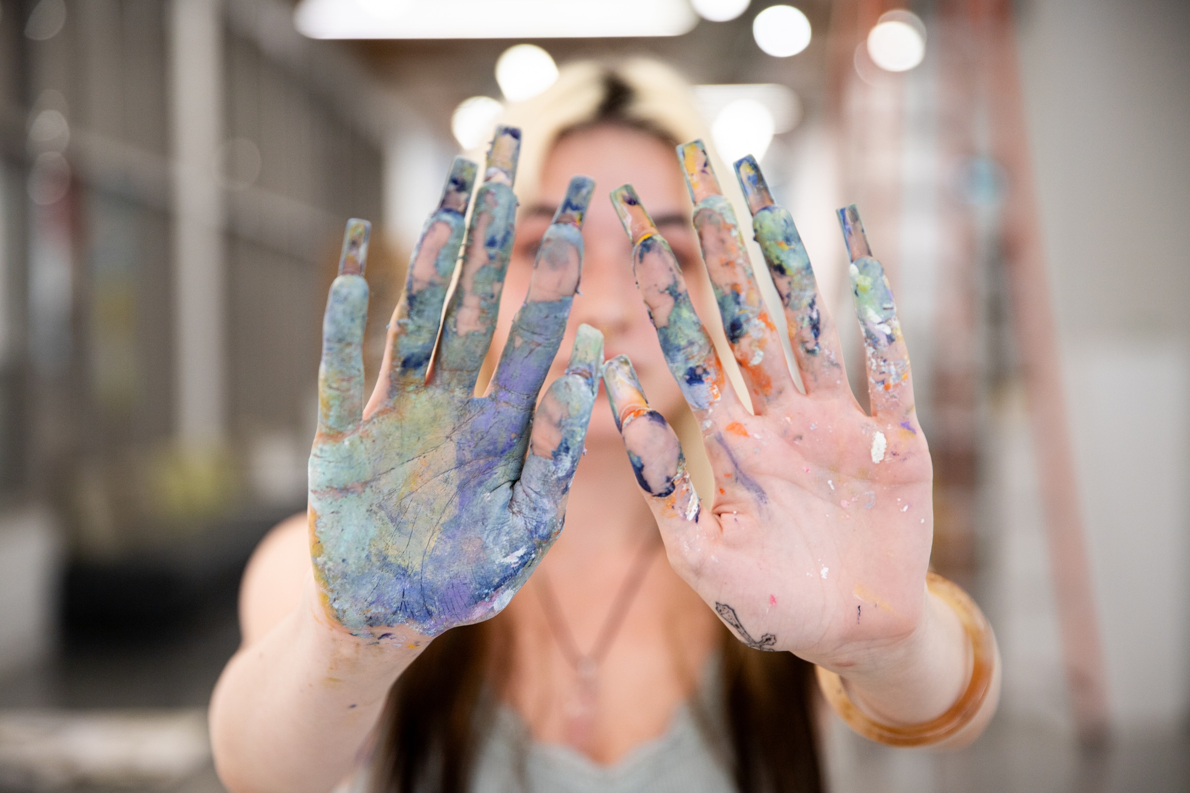 Close-up of a student’s paint-covered hands held forward, with blurred background of an art studio.