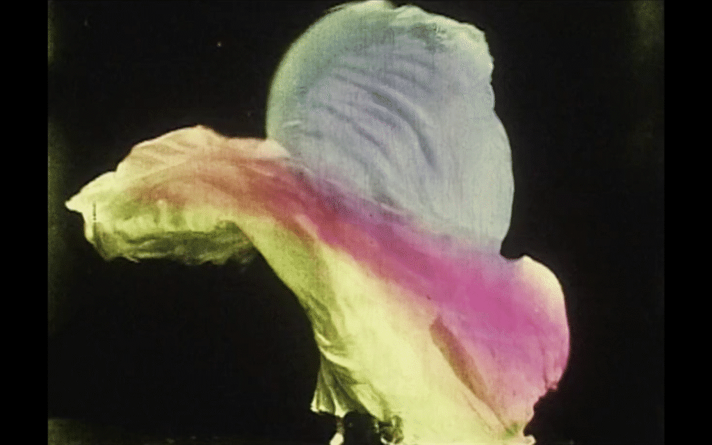 Abstract film still of flowing fabric in green, pink and white tones moving against a dark background.