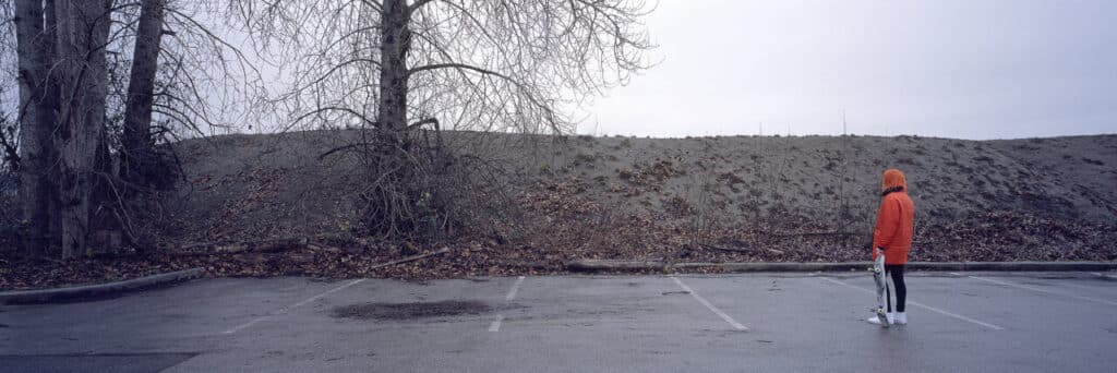 A person in an orange jacket and black pants stands alone in a wet, empty parking lot holding a skateboard. Bare winter trees line the edge of the lot against a pale, overcast sky, creating a quiet, contemplative scene.